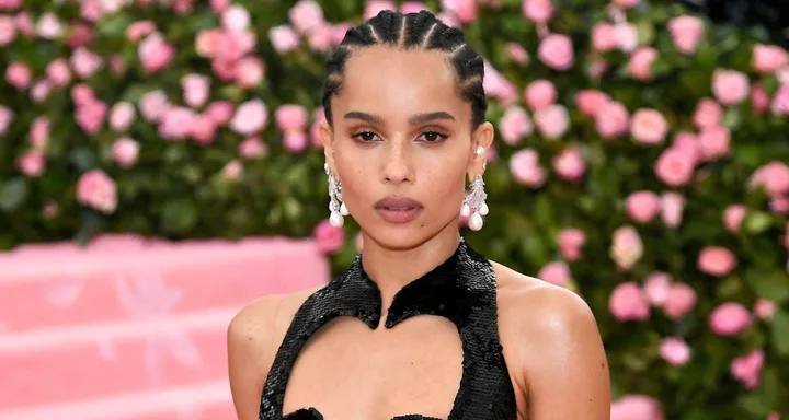 A woman with braided hair and pearl earrings in front of a pink floral backdrop.