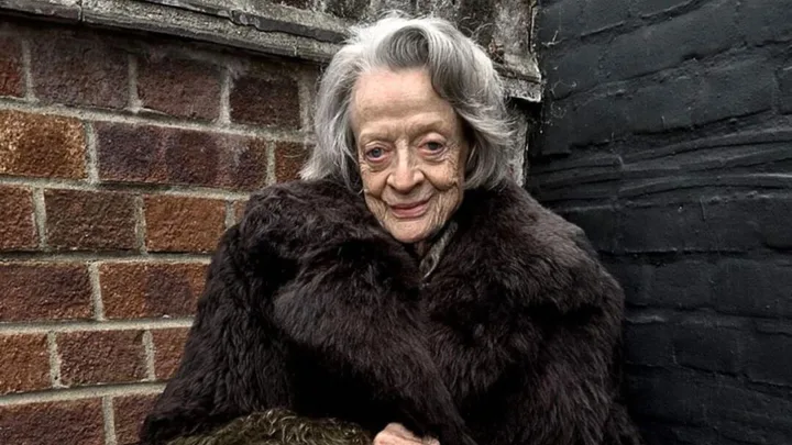 An elderly woman in a dark fur coat standing against a brick wall outdoors.