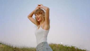 A person in a white tank top and jeans smiles with arms raised against a clear sky background.