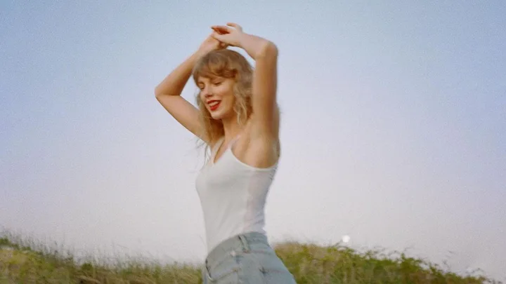 A person in a white tank top and jeans smiles with arms raised against a clear sky background.