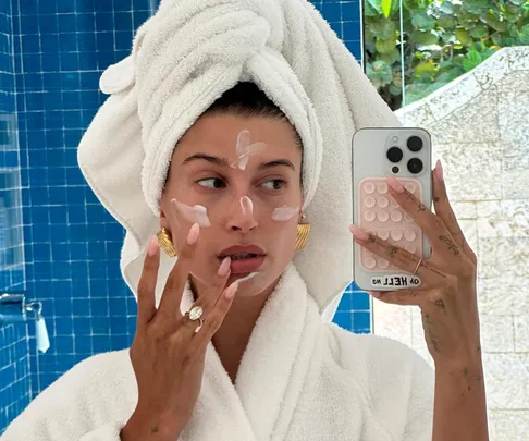 A person in a bathroom wears a towel turban and robe, applying cream to their face while taking a selfie.