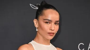 A person with dark hair in a bun, wearing pearl earrings and a light-colored top, in front of a dark background.