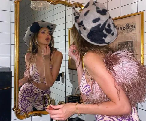 A person in a zebra-pattern outfit and feathered shawl checks makeup in a mirror, wearing a stylish spotted hat.