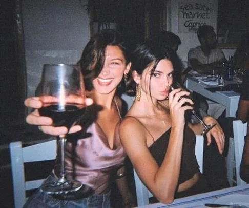 Two women sitting at a restaurant, smiling and holding wine glasses in a lively, dimly lit setting.