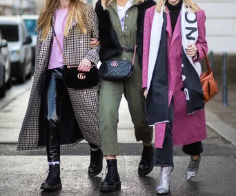 Three stylish women walking in fashionable clothing, featuring long coats, leather pants, and designer handbags.