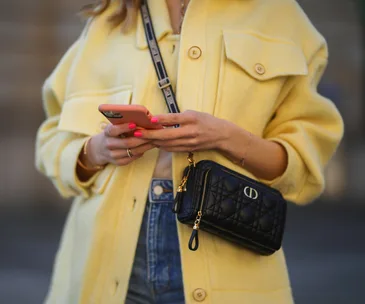 Woman in a yellow coat, holding a smartphone, with a black designer crossbody bag.