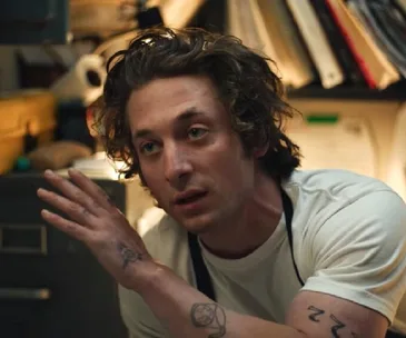 Man with curly hair and tattoos, wearing a white shirt and apron, gesturing in a cluttered room.