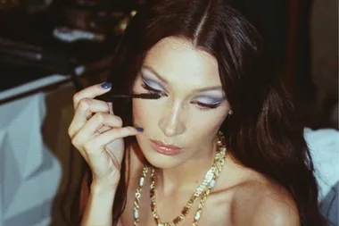 Woman applying mascara with bold eye makeup, wearing a gold necklace.