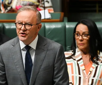 A man speaks in parliament while a woman in glasses sits behind him, listening attentively.