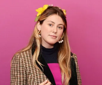 A person with long hair, wearing a checkered jacket and flower headband, stands against a pink background.