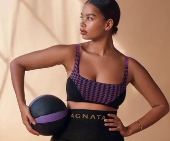 A woman in athletic wear holds a medicine ball, posing confidently against a neutral background.