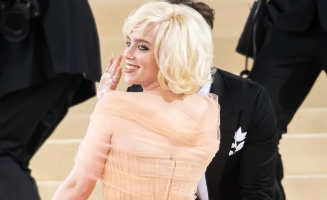 Woman with blonde bob hairstyle and peach gown, smiling and waving on a staircase, with others in formal attire nearby.