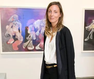 A person standing in front of colorful abstract paintings in a gallery, wearing a black jacket and white blouse.