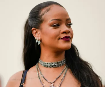 A person with long hair, wearing layered necklaces and earrings, looking to the side, against a neutral background.