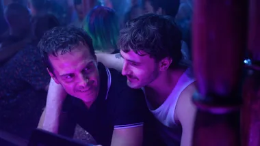 Two men smiling in a crowded nightclub, illuminated by purple light, one with an arm around the other.