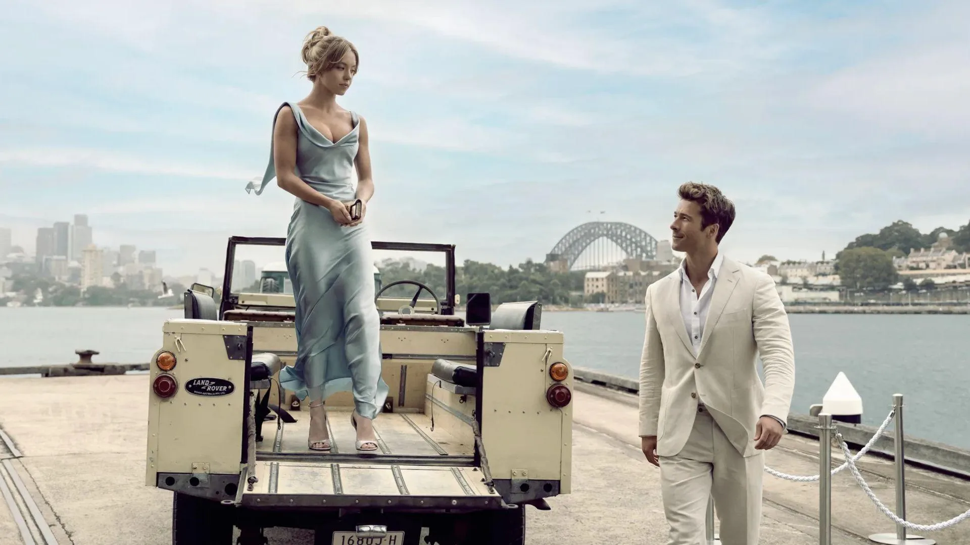 A woman in a light blue dress stands on a vintage jeep beside a man in a white suit, with the Sydney Harbour Bridge in view.