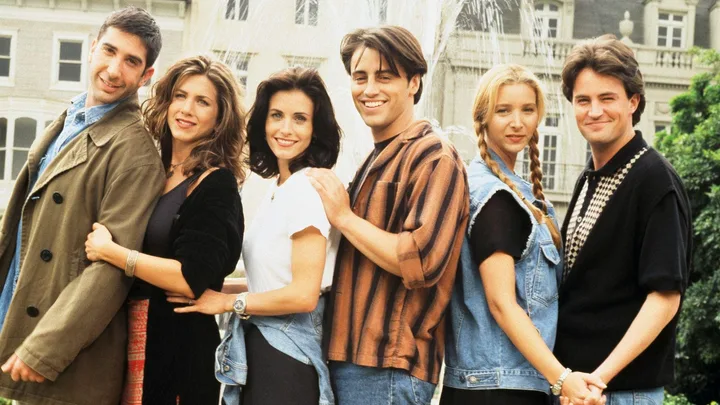 The cast of "Friends" in front of a fountain, smiling and standing in a line outdoors.