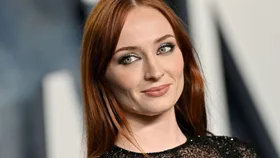 Portrait of a woman with long red hair and a black sequined top, smiling softly with a blurred gray background.