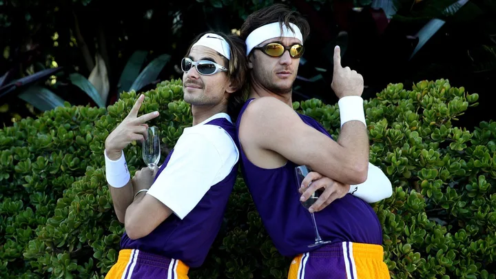 Two men in retro tennis outfits, posing back-to-back with sunglasses and white headbands, holding champagne glasses.