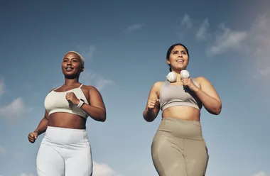 Two women jogging outside in athletic wear, one with headphones around her neck, against a blue sky backdrop.