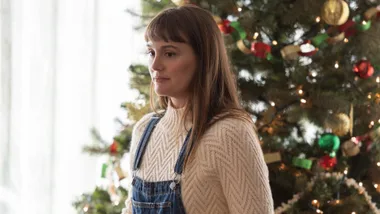 A woman in a cream sweater and denim overalls stands by a decorated Christmas tree, looking thoughtful.