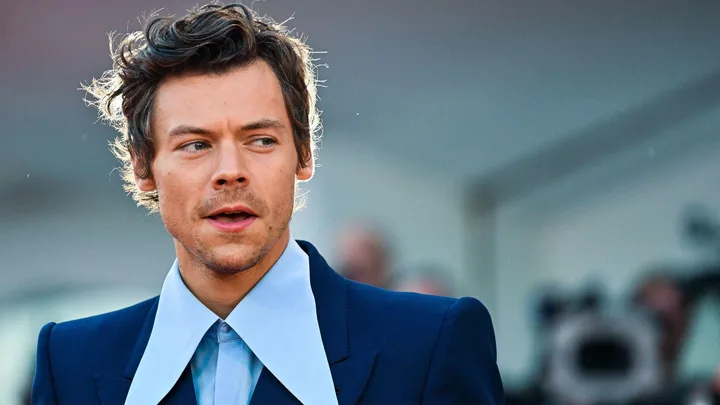 Man in a blue suit with a wide-collared shirt, looking to the side against a blurred background.