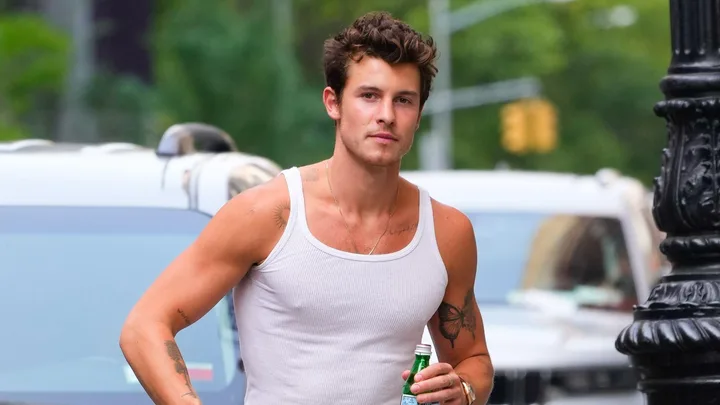 A person in a white tank top holding a drink, walking on a city street with cars in the background.