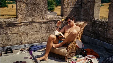 Man lounging in a chair on a rooftop, reading a book and surrounded by binoculars, a badminton racket, and various items.