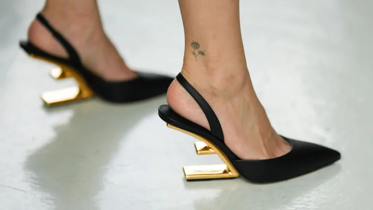 Close-up of black shoes with gold geometric heels on person's feet with a flower tattoo on the ankle.