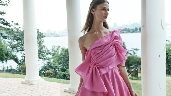 A woman in a pink off-shoulder dress poses outdoors near columns, with trees and a distant city skyline in the background.