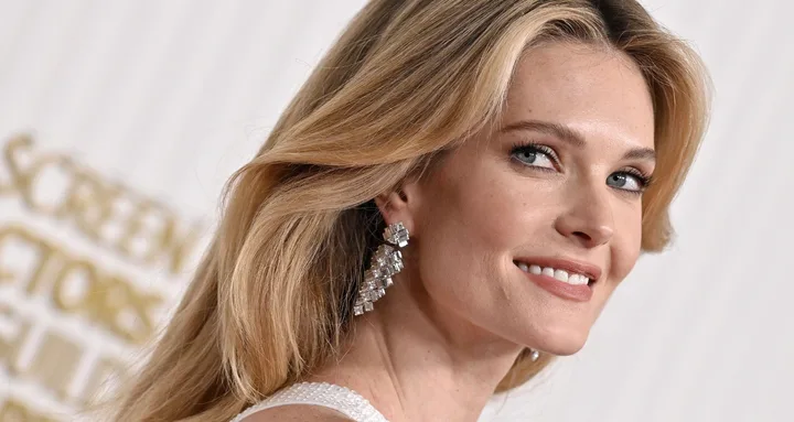 A person with long blonde hair and sparkly earrings smiling at an event, with a blurred background.