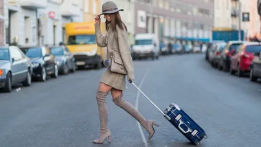 Woman in a hat and boots pulling a blue carry-on luggage on a city street.