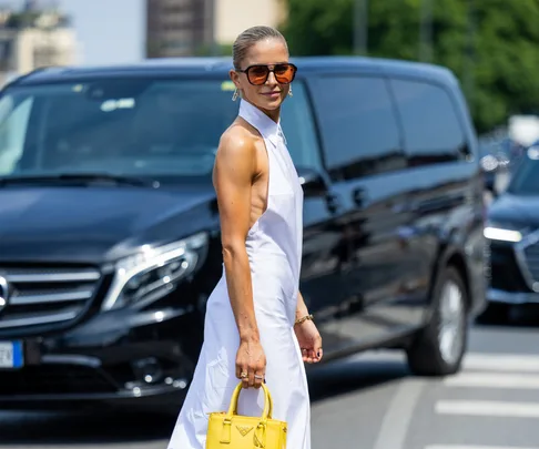 A person in a white dress holding a yellow handbag, standing in front of black cars on a city street.