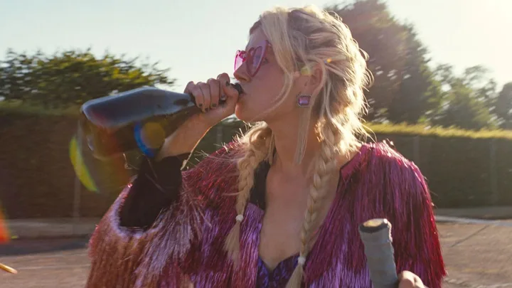 Woman with braided hair and heart-shaped sunglasses drinks from a champagne bottle, wearing a shiny pink jacket.