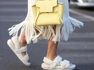 Woman wearing white fluffy sandals, a white fringed dress, and carrying a yellow bag, walking on a street.