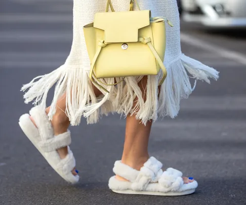 Woman wearing white fluffy sandals, a white fringed dress, and carrying a yellow bag, walking on a street.