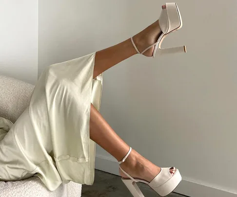 Woman lying back with legs up, wearing ivory platform heels and a satin dress, against a plain backdrop.