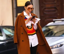 A woman in a brown coat and colorful sweater looks at her phone while walking on a city street.