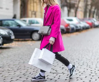 Woman in pink coat walking down cobblestone street with shopping bags and crossbody bag.