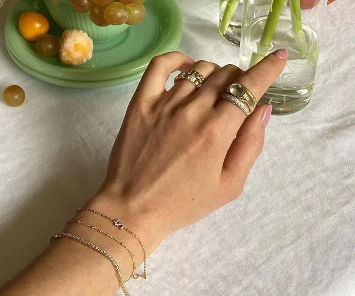 A hand adorned with rings and bracelets touching a glass vase filled with water on a table set with fruits.