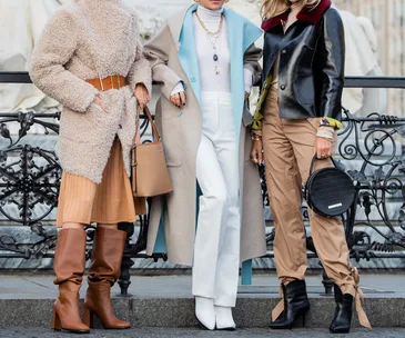 Three fashion-forward women in stylish coats and boots stand confidently on the street.