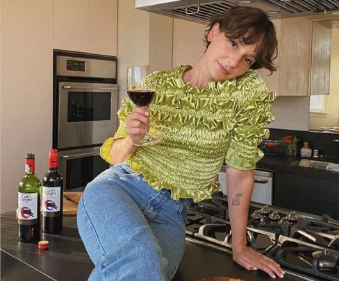 Person in a green top holding a wine glass, sitting on a kitchen counter with two wine bottles visible.