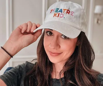 A woman wearing a hat that says "Theatre Kid" poses with a slight smile indoors.