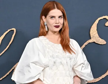 A person with long red hair, dark lipstick, wearing a white textured dress against a dark background with gold accents.