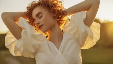 A redhead girl smiling in the sunlight wearing a puff sleeved white dress.