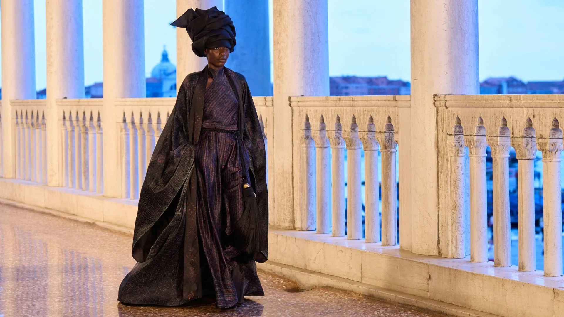 Model in an elegant dark outfit and headdress walks on a balcony with white pillars against a blue sky background.