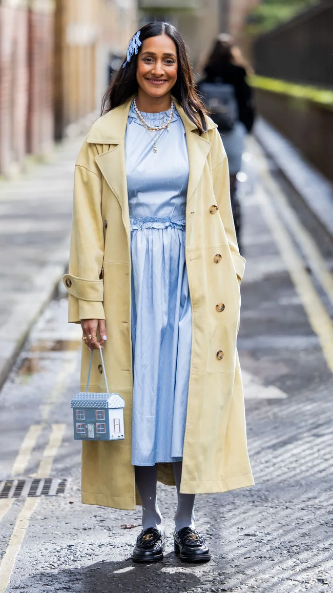 Zeena Shah wears blue bows in hair, blue dress, pearl necklace, beige coat, Chanel loafers, tights, blue bag outside Eudon Choi during London Fashion Week February 2024 on February 18, 2024 in London, England.