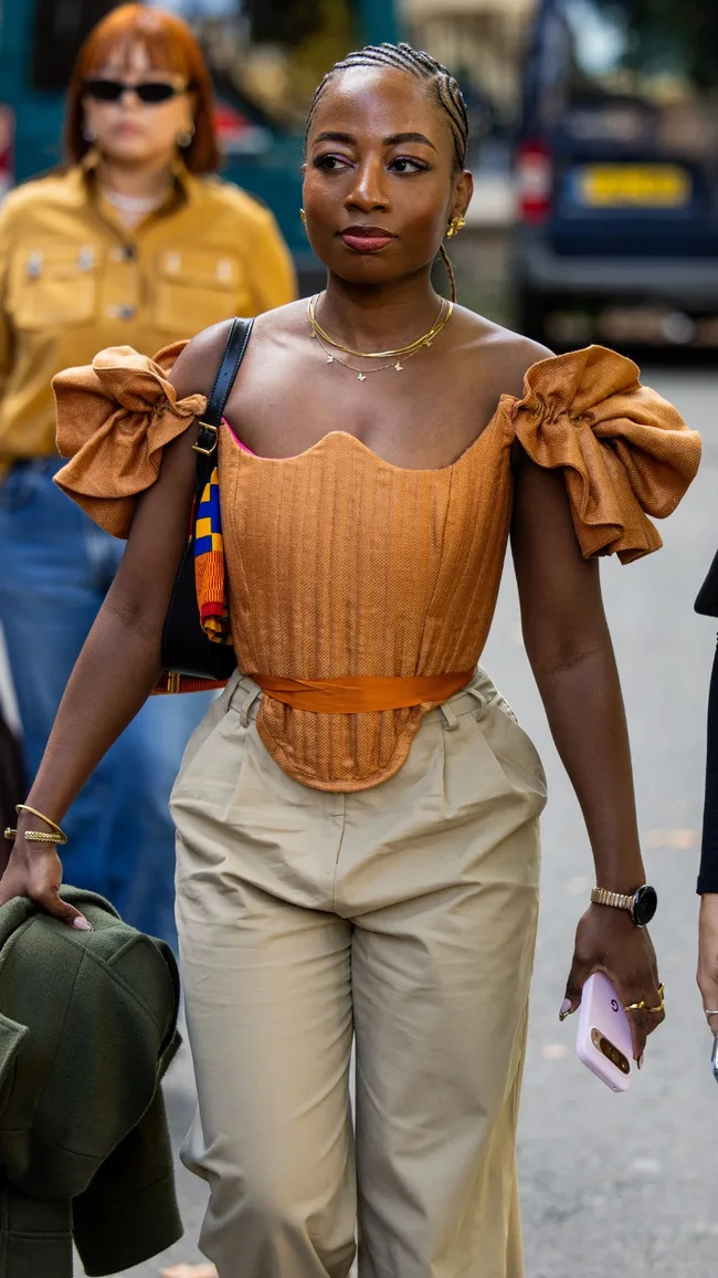 A guest wears rust brown off shoulder top, beige pants outside Ahluwalia during London Fashion Week September 2024 on September 14, 2024 in London, England.