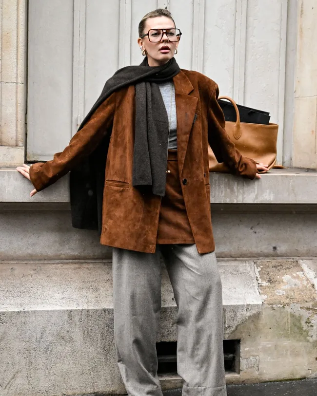 woman poses against wall at paris fashion week wearing brown suede jacked grey trousers and a scarf