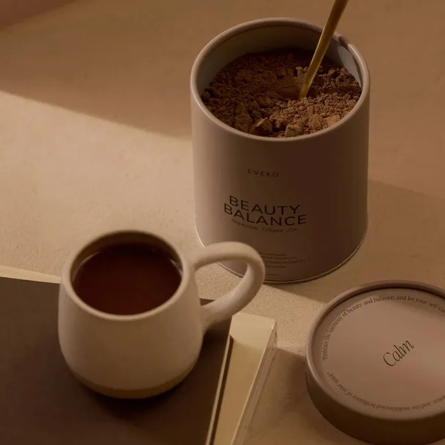A mug of coffee next to an open container of Eveko Beauty Balance powder with a wooden spoon inside.